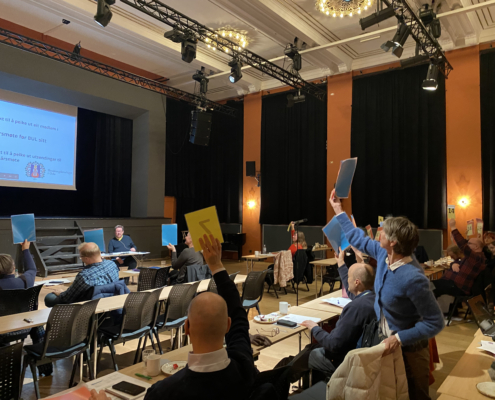 Møte i stor sal. Menn og kvinner viser stemmeseddel.